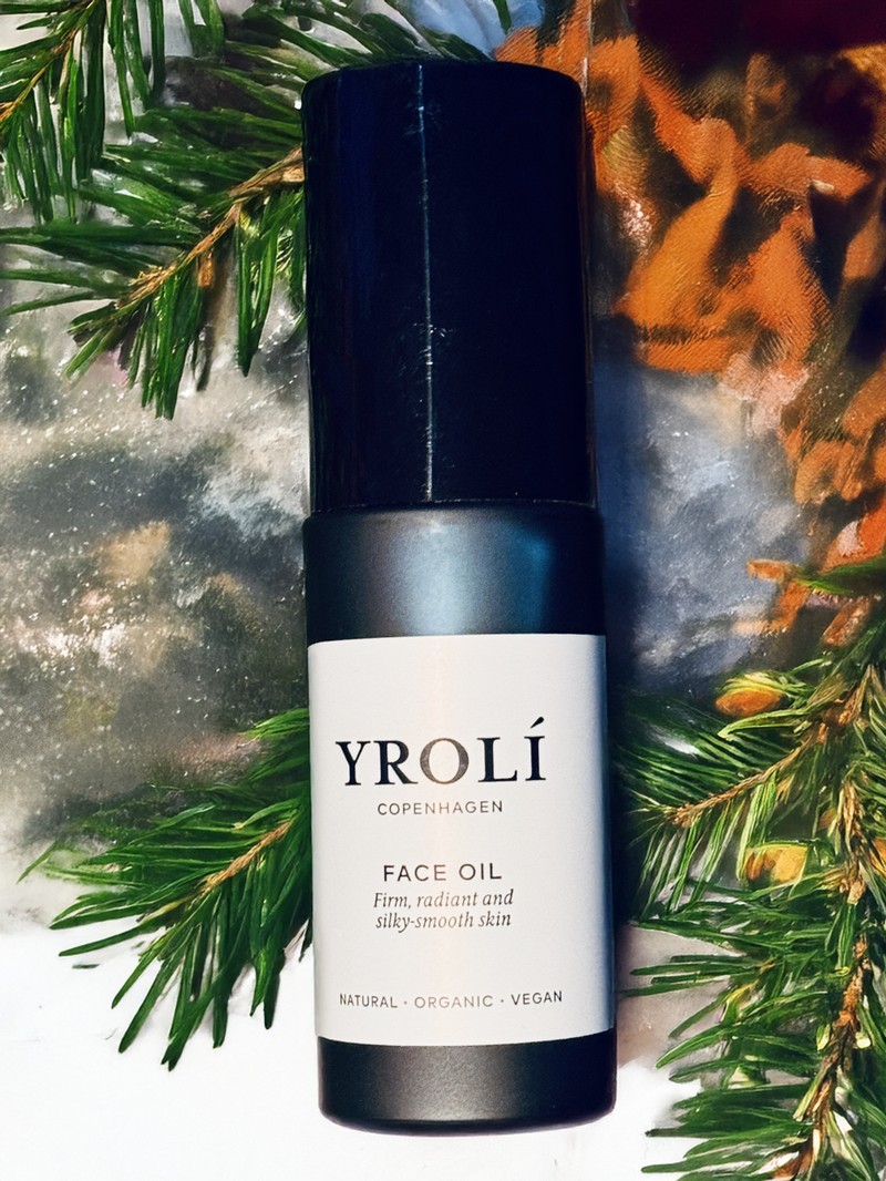 Eine schwarze Flasche Gesichtspflege-Öl der skandinavischen Marke Yrolí liegt auf frischen, dunkelgrünen Tannenzweigen. Auf dem hellen Etikett der mattschwarzen Flasche steht „YROLÍ Copenhagen – Face Oil. Firm, radiant and silky-smooth skin. Natural, Organic, Vegan“. Das Licht spiegelt sich leicht auf dem Deckel, während die umgebenden Zweige einen weihnachtlichen, natürlichen Rahmen bilden.