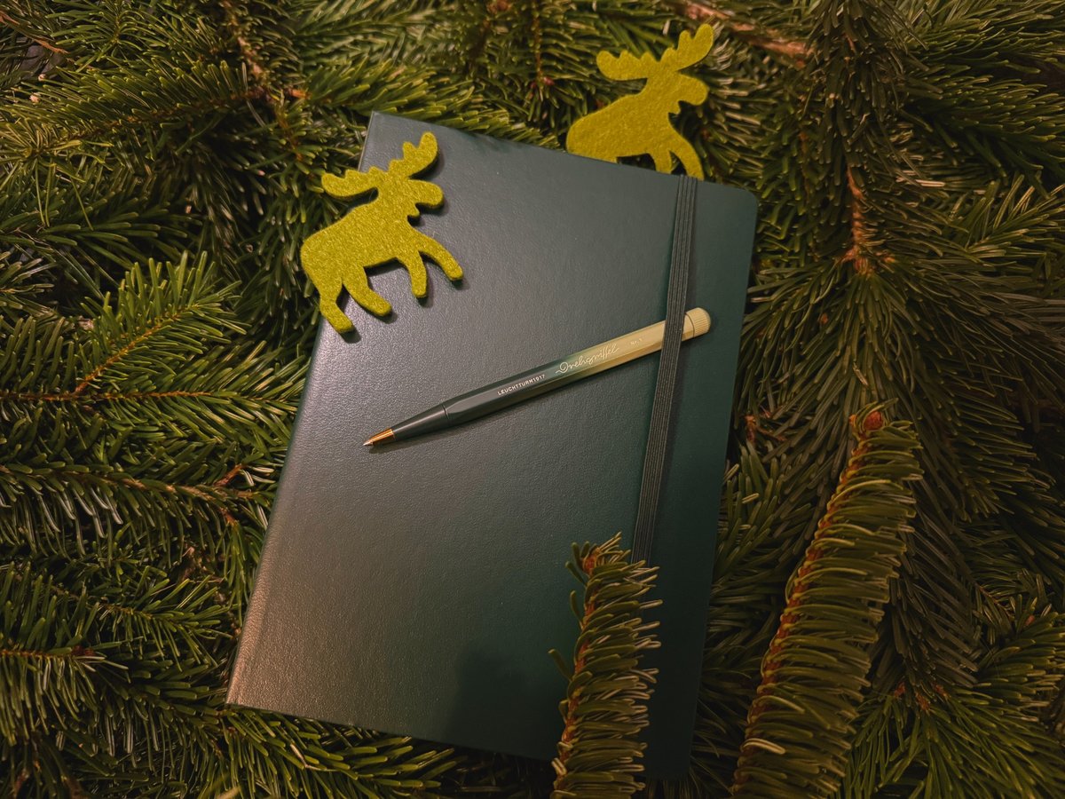 Ein dunkelgrünes Notizbuch von Leuchtturm1917 liegt zwischen Tannenzweigen, darauf ein grüner Drehgriffel; zwei kleine Filz-Elche bilden eine dekorative Umrahmung.