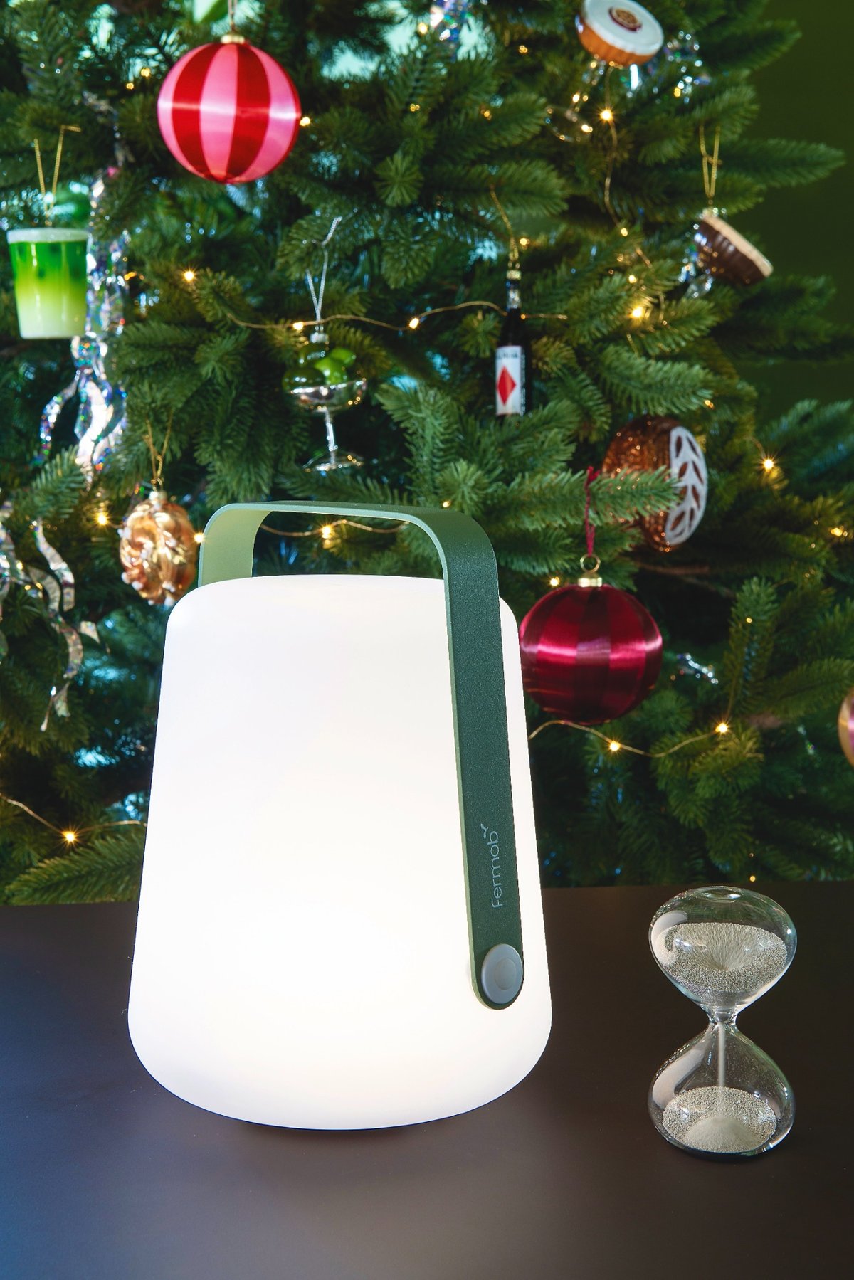 Eine mobile LED-Lampe von Fermob steht auf einem Tisch vor einem geschmückten Weihnachtsbaum. Daneben liegt eine gläserne Sanduhr.