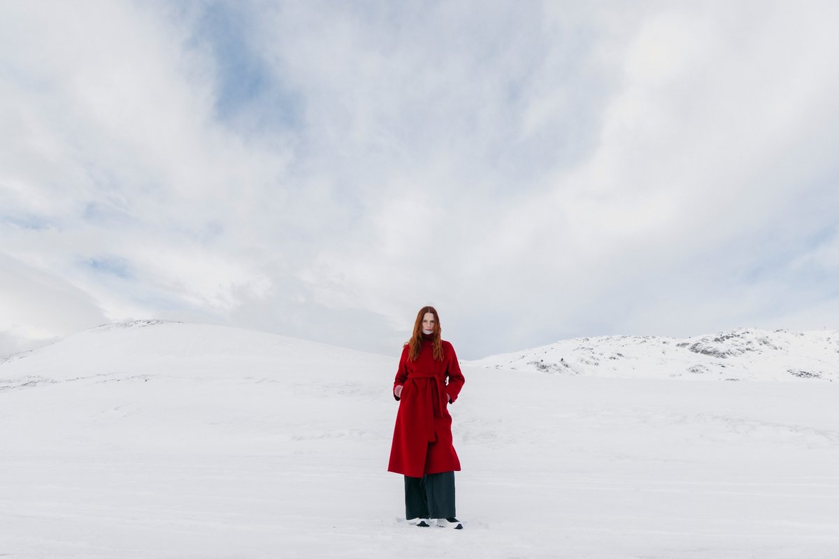 Eine Frau mit langen roten Haaren steht in einem roten Wollmantel in einer verschneiten Landschaft mit Hügeln unter bewölktem Himmel.