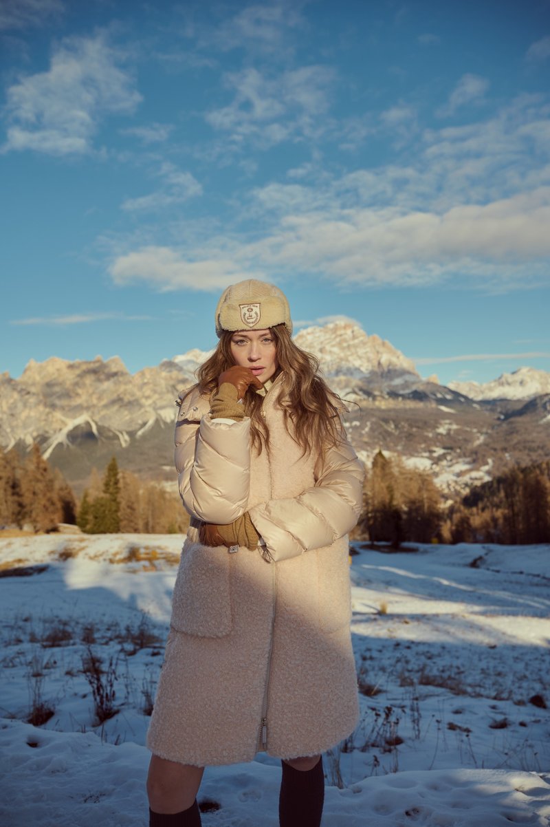 Person trägt einen hellen, knielangen Mantel mit Stehkragen und Mütze in einer verschneiten Berglandschaft.