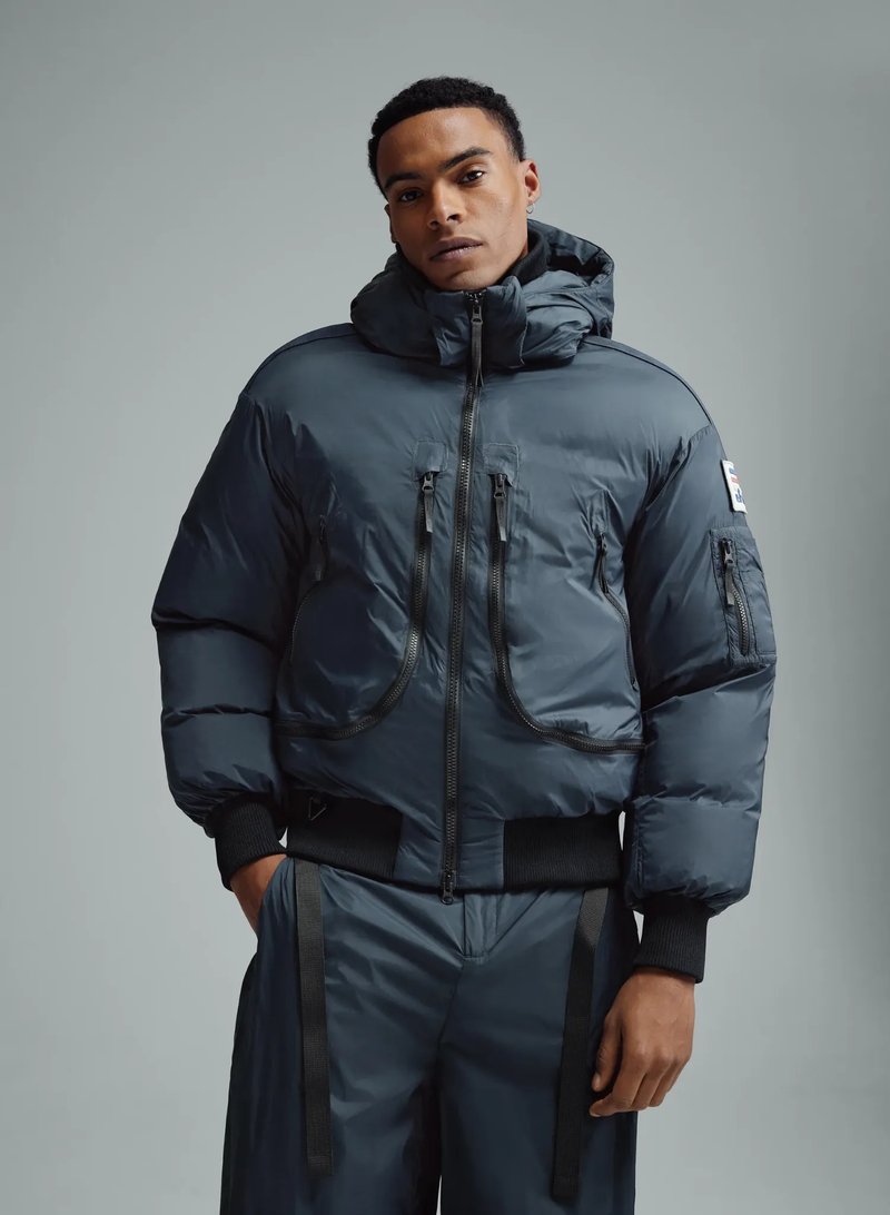 Eine männliche Person trägt eine dunkelblaue, wattierte Bomberjacke mit Kapuze, Frontreißverschluss und mehreren Reißverschlusstaschen vor neutralem Hintergrund.