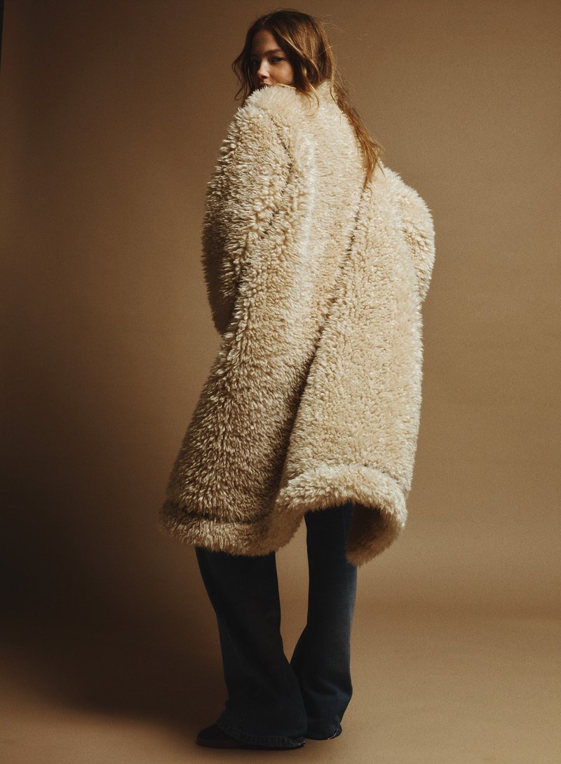 Person trägt einen hellen, knielangen Kunstpelzmantel mit voluminöser Oberfläche vor einfarbigem Hintergrund.