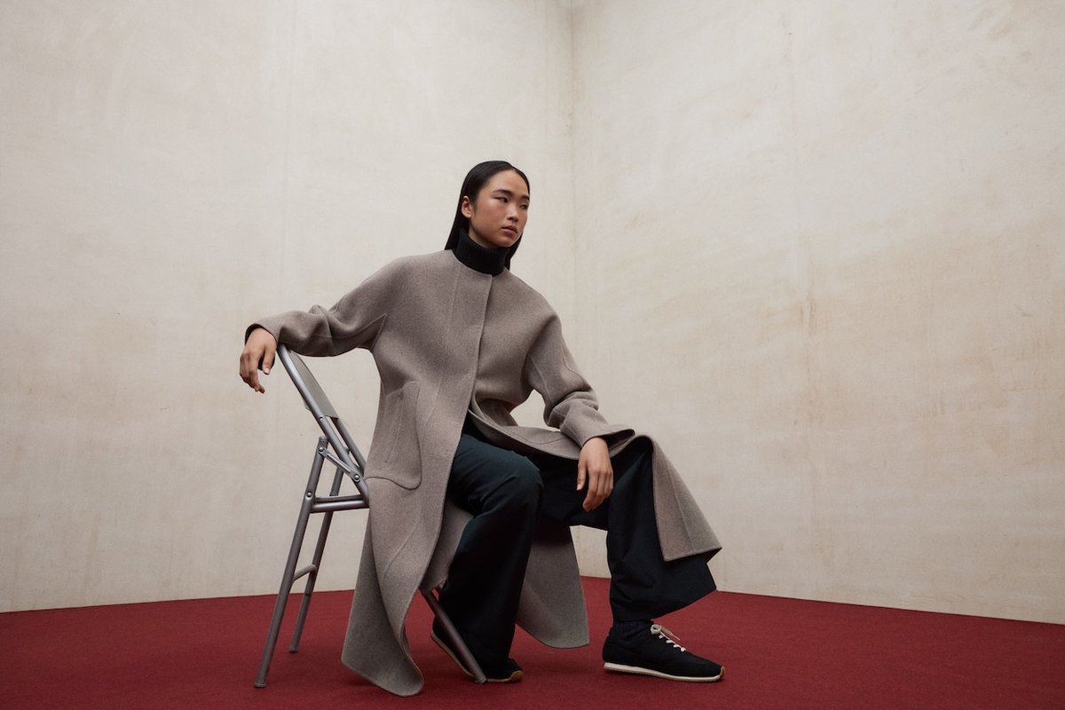 Person sitzt auf einem Stuhl und trägt einen langen, grauen Mantel mit Rundhalsausschnitt und verdecktem Verschluss vor neutralem Hintergrund.