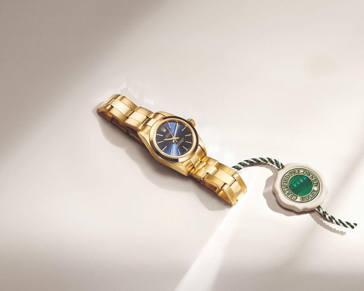 Eine goldene Rolex Oyster Perpetual mit blauem Zifferblatt und Certified-Pre-Owned-Siegel auf hellem Untergrund.