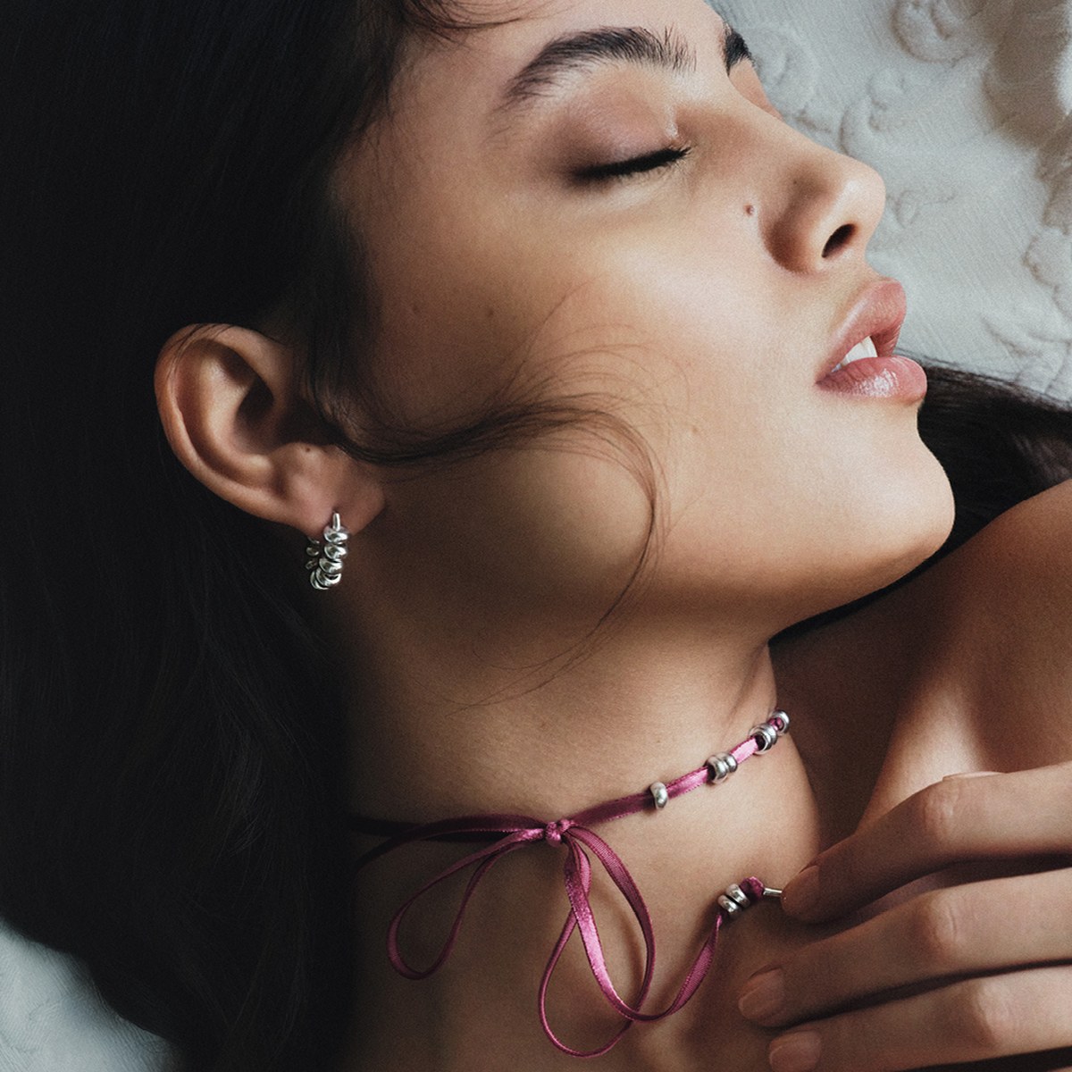 Close-up-Foto: Model liegt seitlich auf hellem Stoff, trägt rosafarbenes Samtband mit kleinen silbernen Elementen sowie Creolen aus Silber.