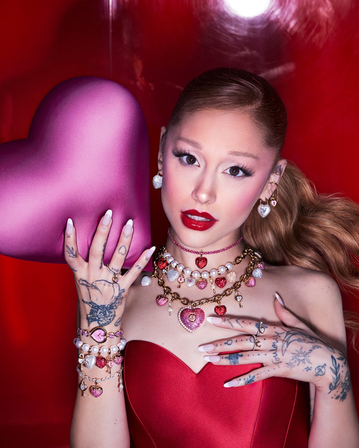 Ariana Grande vor rotem Hintergrund, hält ein pinkfarbenes Herzobjekt und trägt mehrere Halsketten, Armbänder, Ringe und Ohrringe mit Herzmotiven.