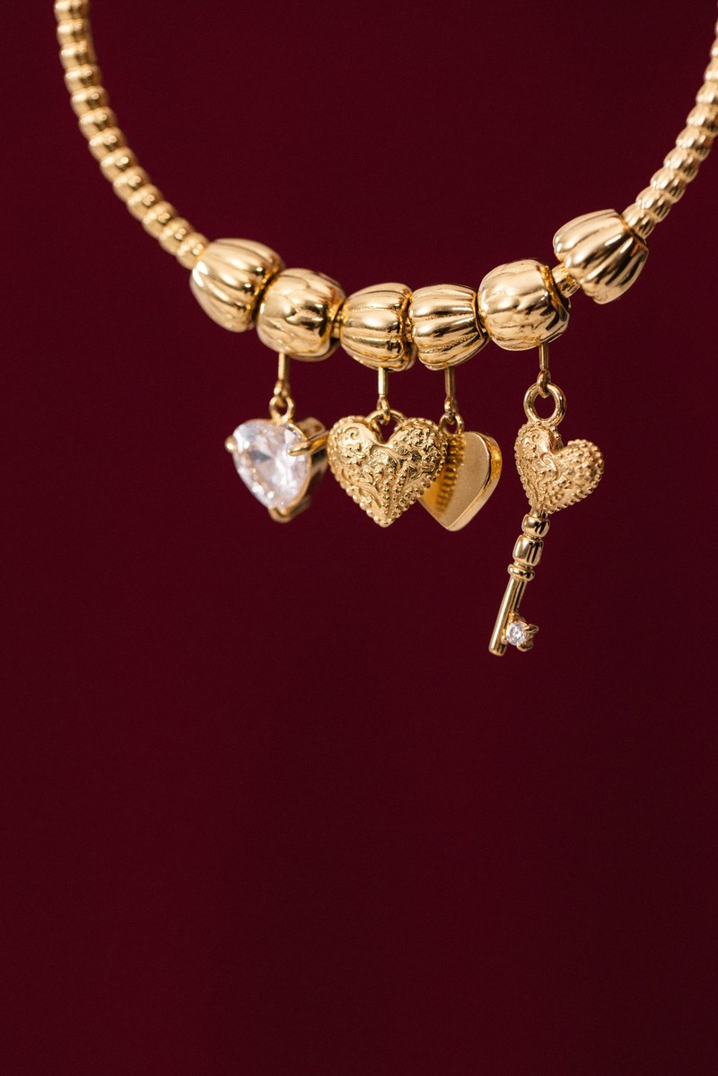 Detailaufnahme eines goldfarbenen Armreifs mit mehreren herzförmigen Charms und einem Schlüsselanhänger vor bordeauxfarbenem Hintergrund. Die Charms sind beweglich am Armreif befestigt.