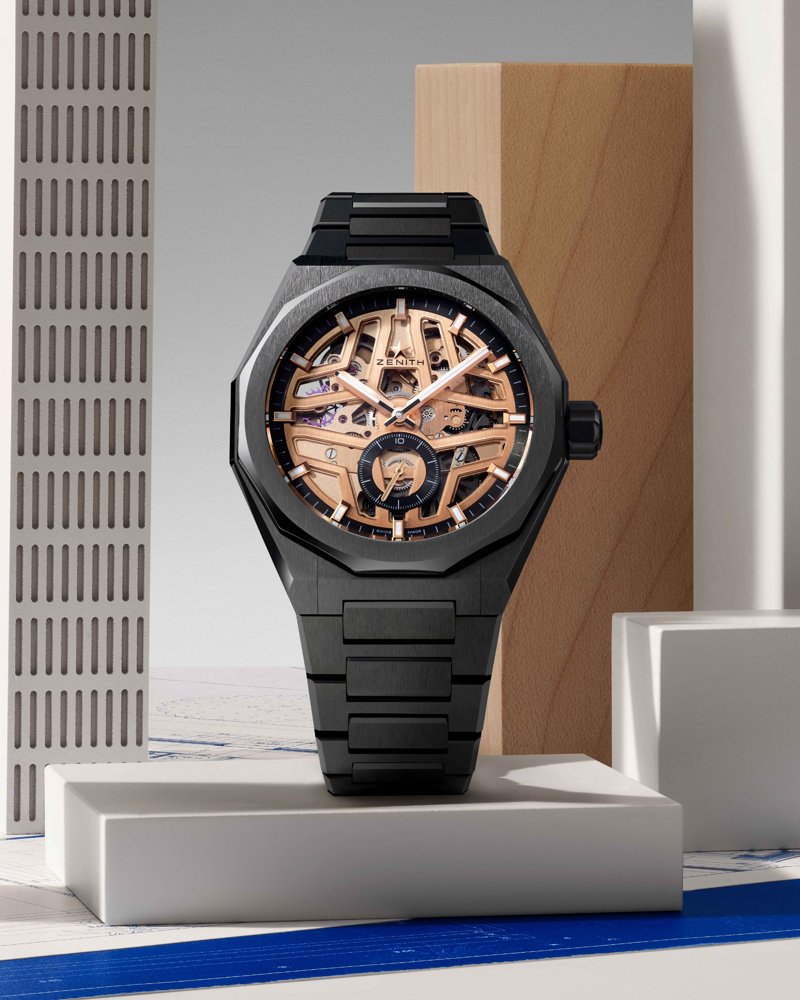 substantial-times-architektur-am-handgelenk-03 Aufnahme einer skelettierten Uhr mit schwarzem Gehäude und sichtbarem Uhrwerk mit goldfarbenen Komponenten, im Hintergrund Dekoration einer Skyline