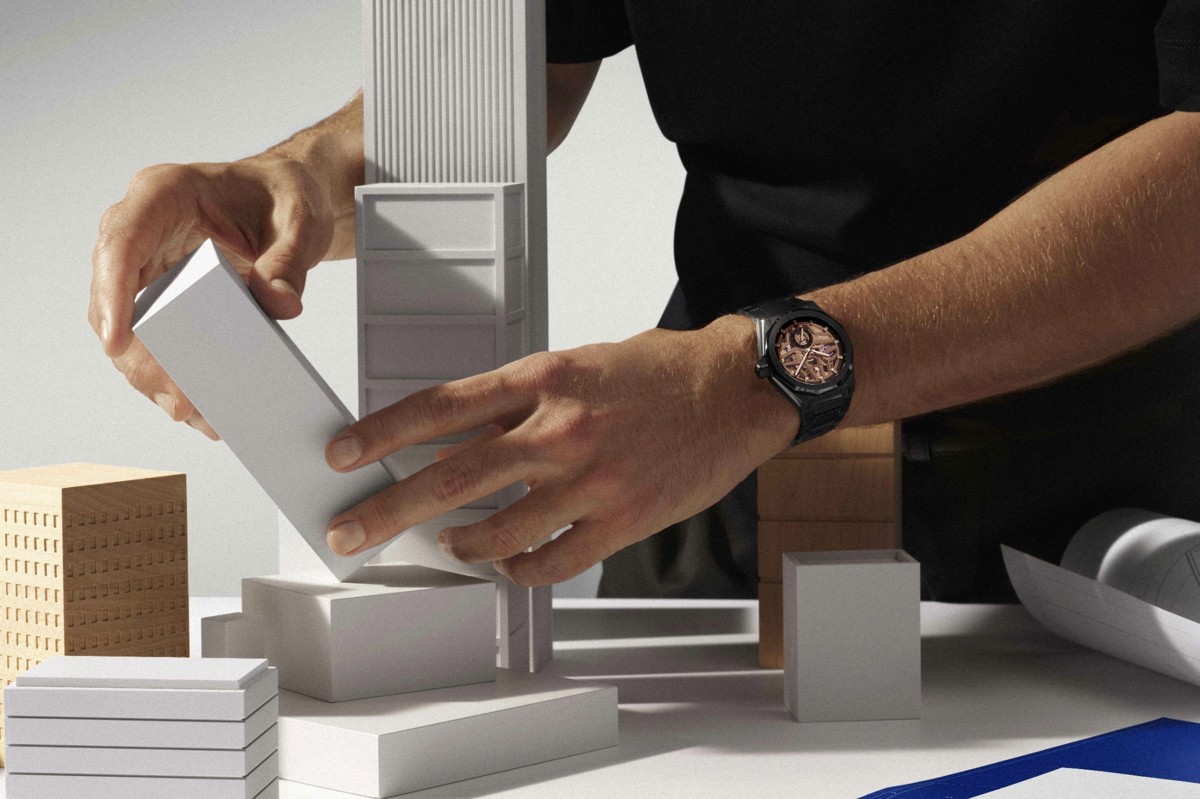 substantial-times-architektur-am-handgelenk-hero-02 Alt-Text: Arm mit schwarzer skelettierter Armbanduhr neben weißen Architekturmodellen