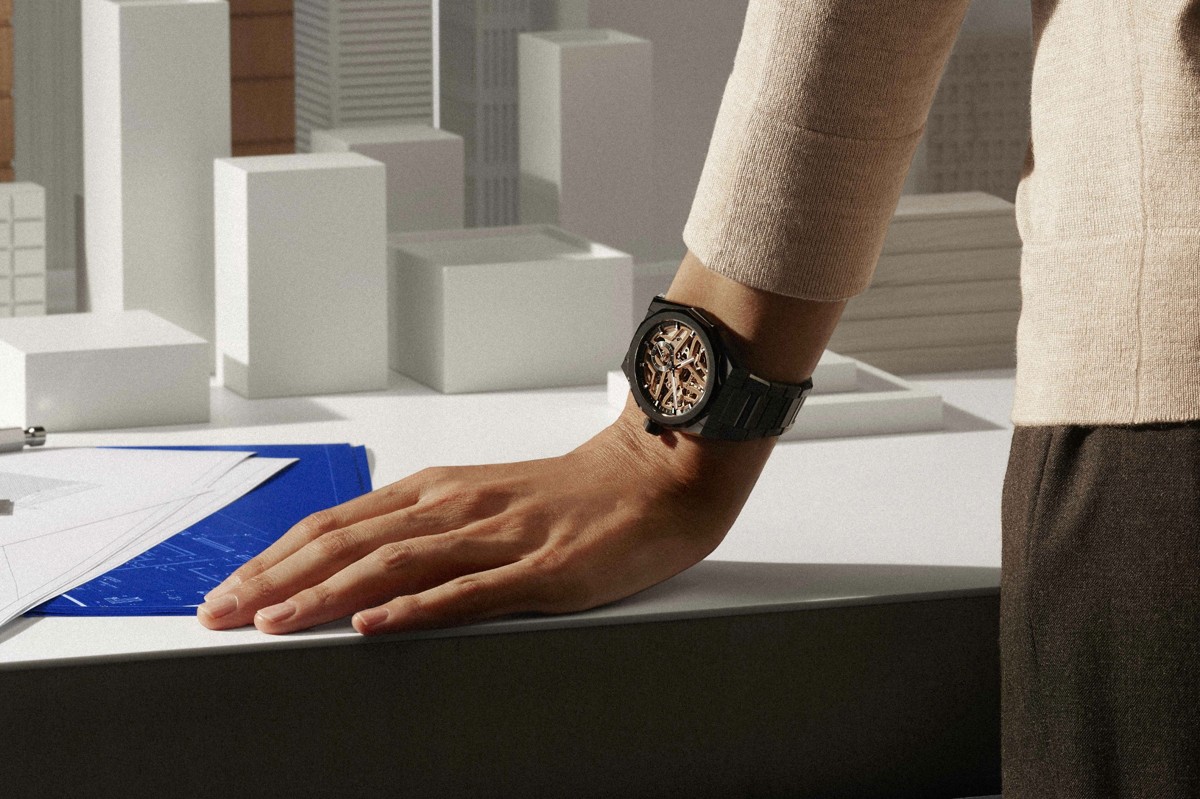 Hand mit schwarzer skelettierter Armbanduhr über Architekturmodellen und Bauplänen