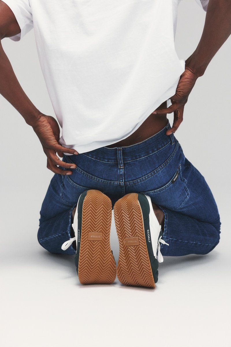 substantial-times-rebellion-der-revoluzzer-ecoalf-denim-02 Männliches Model im Anschnitt, Rückenansicht, er kniet auf dem Boden, trägt dunkle Jeans und Sneaker, Blick auf Schuhsohlen