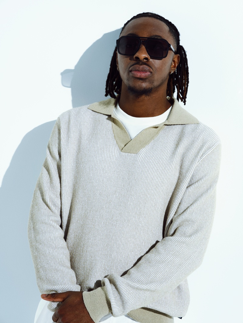 Mann mit geflochtenem Haar trägt eine schwarze, eckige Sonnenbrille und sitzt in hellem Strickpullover vor weißem Hintergrund