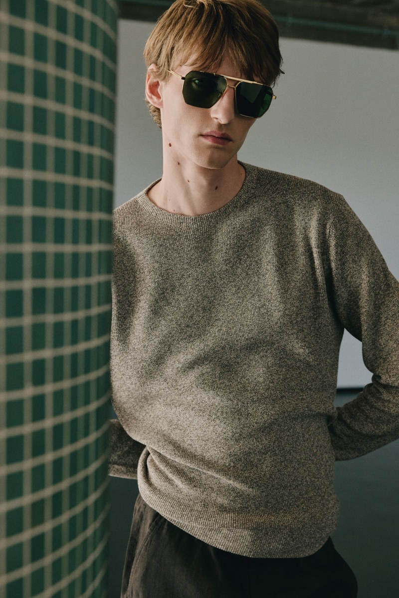 Männliches Model mit goldfarbener, geometrischer Sonnenbrille mit grün getönten Gläsern vor gekachelter Wand