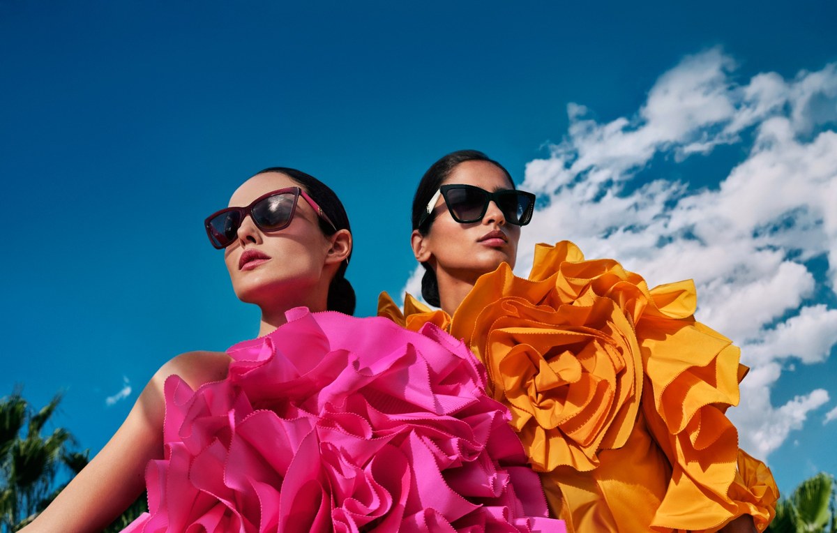 Zwei Models in pinken und orangefarbenen Volantkleidern tragen Cat-Eye-Sonnenbrillen vor blauem Himmel
