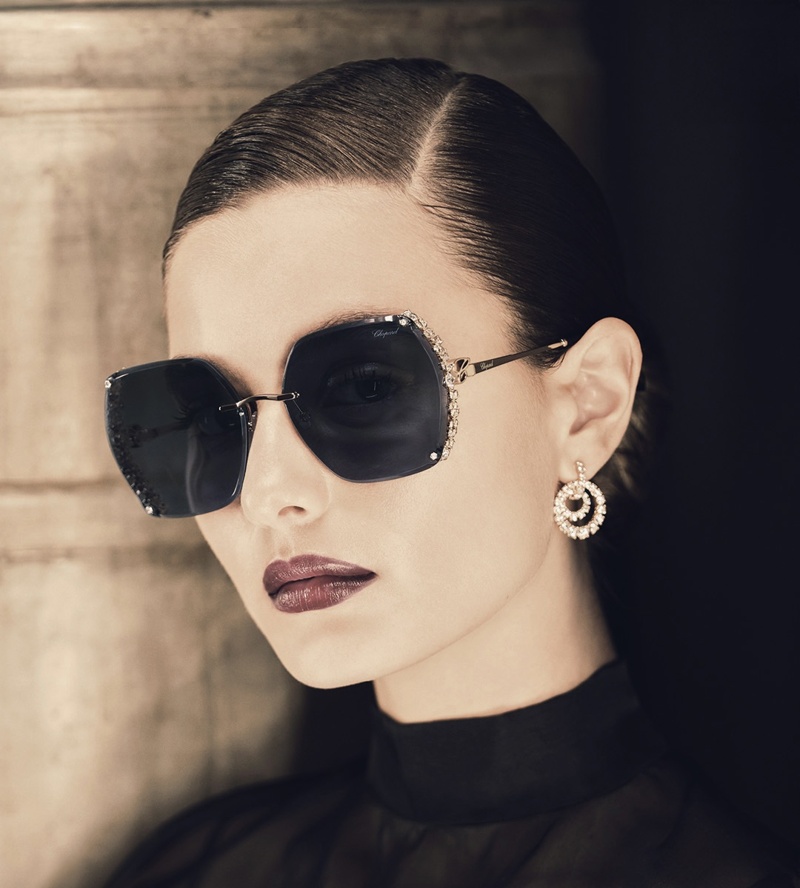 Frau mit streng zurückgekämmtem Haar trägt große Sonnenbrille mit dunklen Gläsern und goldfarbenem Metallrahmen mit seitlichen Kristallen