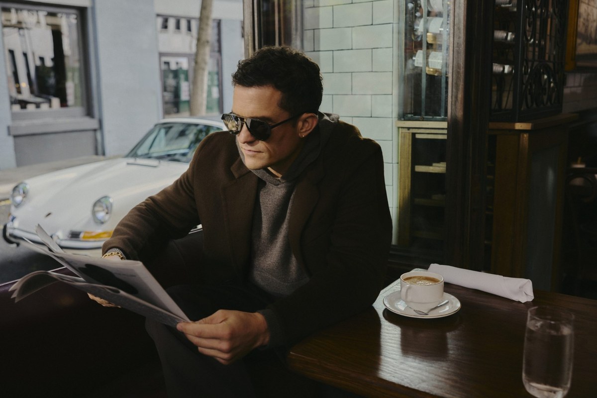 Orlando Bloom sitzt in einem Café und trägt eine Aviator-Sonnenbrille von Porsche Design mit Metallrahmen