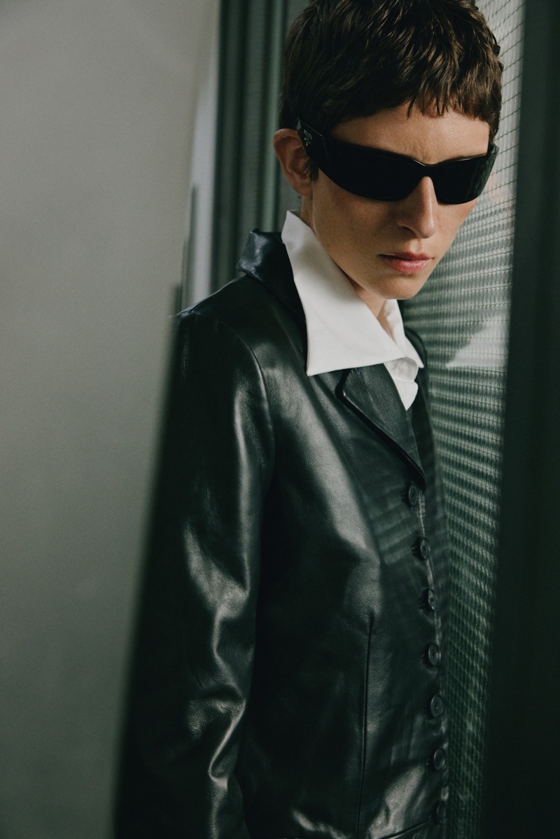 Model mit schwarzer, schmaler Sonnenbrille und Lederjacke in Innenraum mit Fenster