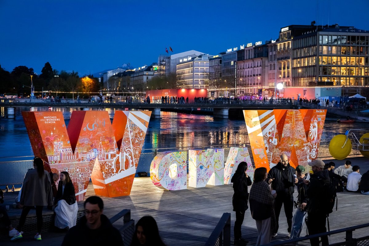 Beleuchtete Installation mit Schriftzug von Watches & Wonders am Seeufer in Genf bei Nacht, umgeben von Besuchern