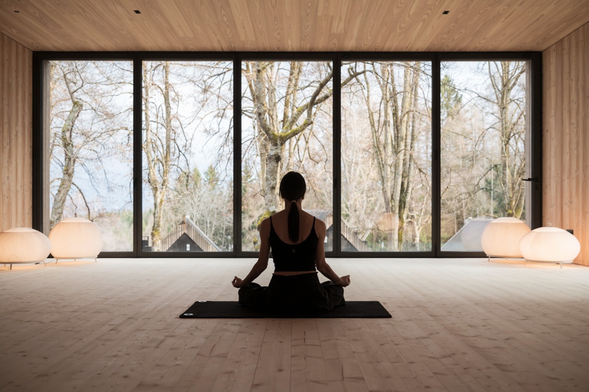 Frau sitzt meditierend auf einer Yogamatte in einem offenen Pavillon mit großer Glasfront und Blick in den Wald
