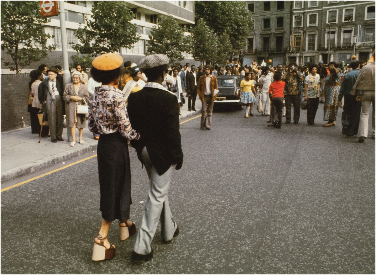 Zwei elegant gekleidete Menschen gehen während des Notting Hill Carnival eine Straße entlang. Im Hintergrund stehen und laufen weitere Passanten