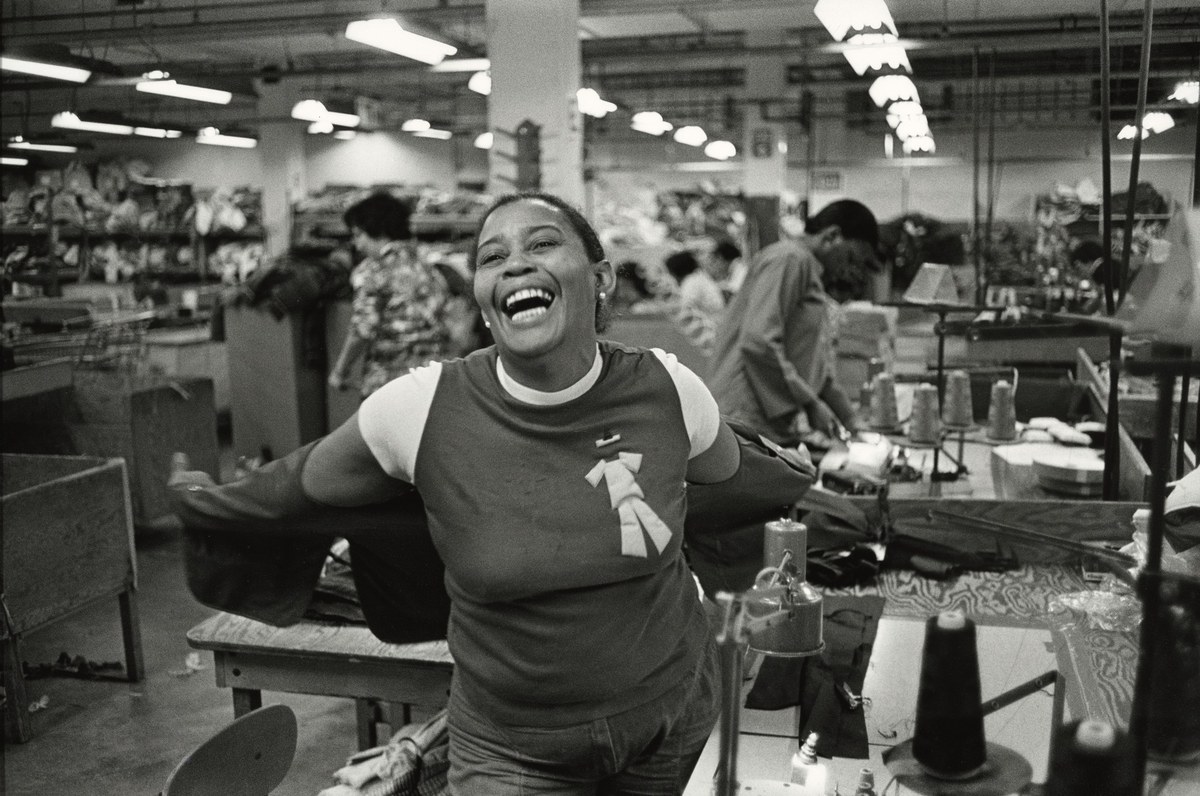 Schwarzweißfotografie einer lachenden afroamerikanischen Frau an ihrem Arbeitsplatz in einer Textilfabrik.