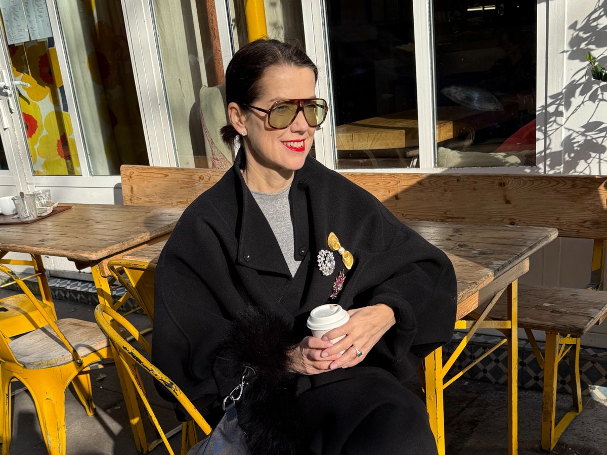 Frau mit Sonnenbrille sitzt an einem Holztisch im Freien, trägt einen schwarzen Mantel mit Broschen und hält einen Coffee-to-go-Becher, im Hintergrund gelbe Metallstühle und Fensterfront.