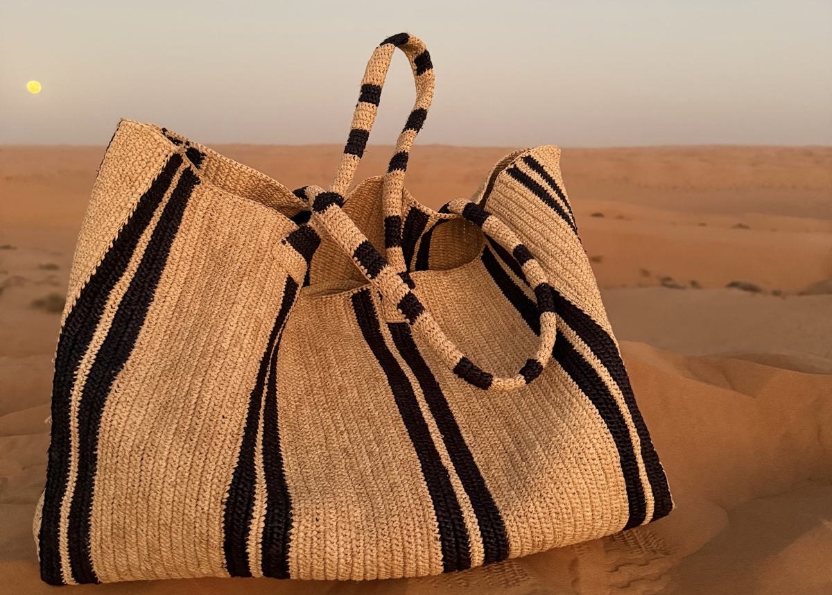 Große Raffia-Tasche mit schwarzen Streifen steht in einer sandigen Dünenlandschaft, aufgenommen bei warmem Licht, mit sichtbaren Fußspuren im Sand.