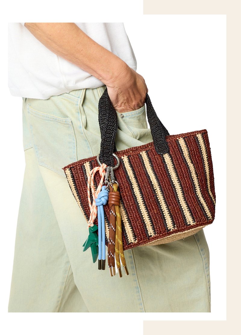Kleine gestreifte Raffia-Tasche in Braun- und Naturtönen wird in der Hand getragen, daran befestigt mehrere bunte Charms aus Kordeln und Anhängern.