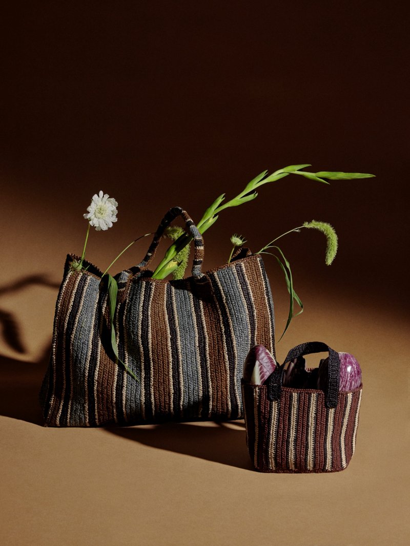 Zwei gestreifte Raffia-Taschen in Braun- und Naturtönen stehen auf braunem Untergrund, eine größere mit eingesetzten Blumen, daneben eine kleinere Tasche mit Gemüse.