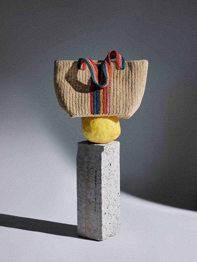 Kleine Raffia-Tasche mit bunten Streifen steht auf einem Steinblock, darunter eine große Zitrone, vor neutralem Hintergrund.