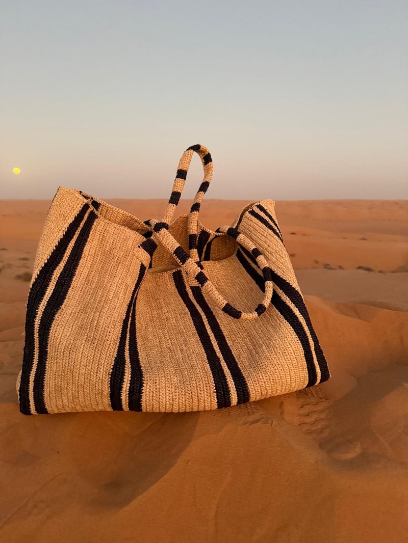 Große Raffia-Tasche mit schwarzen Streifen steht in einer sandigen Dünenlandschaft, aufgenommen bei warmem Licht, mit sichtbaren Fußspuren im Sand.