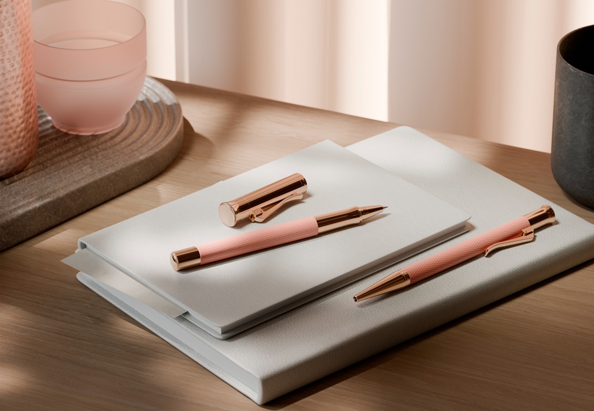Tintenroller und Kugelschreiber in Rosé mit guillochierter Oberfläche liegen auf einem Notizbuch auf einem Holzschreibtisch