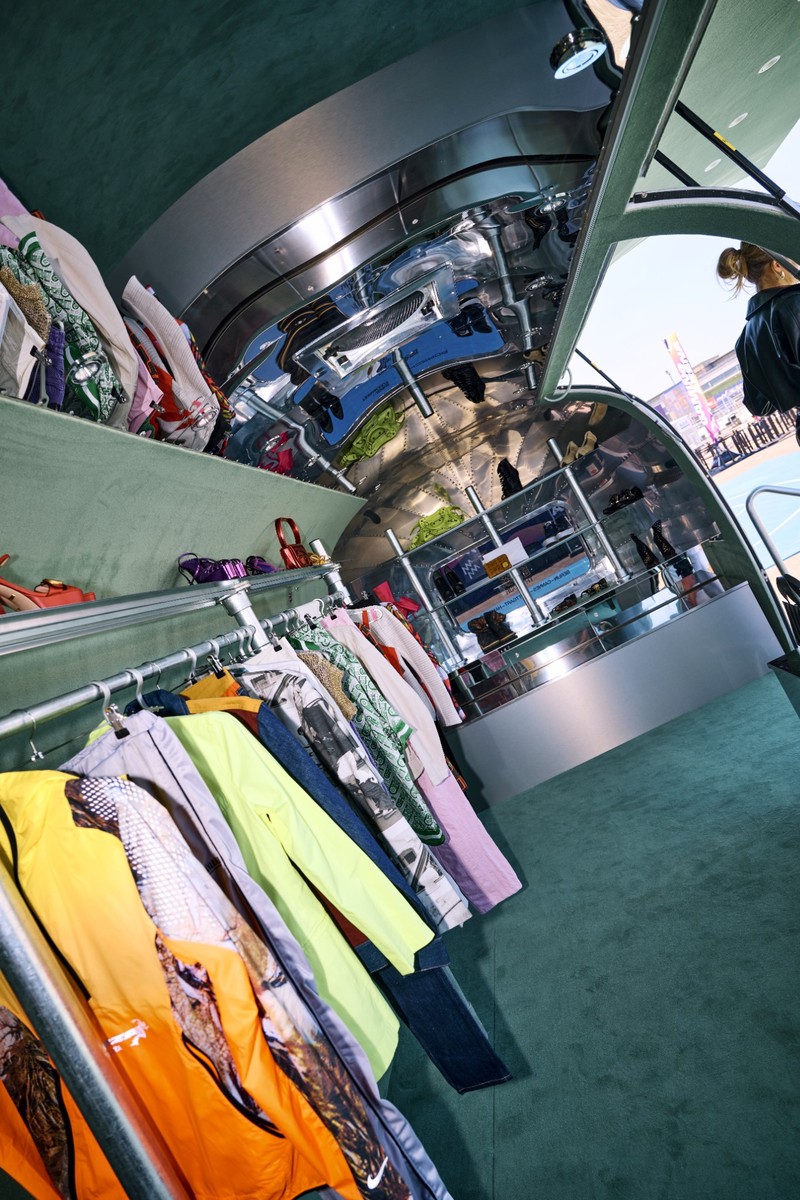 Blick in den Innenraum der Airstream-Boutique: Im Vordergrund eine Kleiderstange mit bunten Designerstücken in Orange, Gelb und Grün, dahinter spiegelt das polierte Aluminium der Wölbung die gesamte Kollektion. Im Hintergrund eine Verkaufstheke und der Ausblick auf das sonnige Tempelhofer Feld.