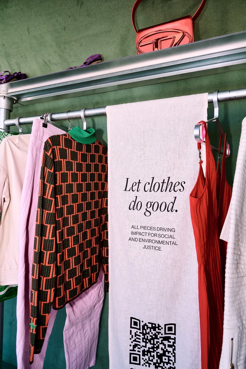 Eine Kleiderstange vor grünem Hintergrund mit bunten Designerstücken – darunter ein gemusterter Rollkragenpullover und ein rotes Trägerkleid. In der Mitte hängt ein helles Leinenbanner mit der Aufschrift Let clothes do good. All pieces driving impact for social and environmental justice, sowie einem QR-Code.