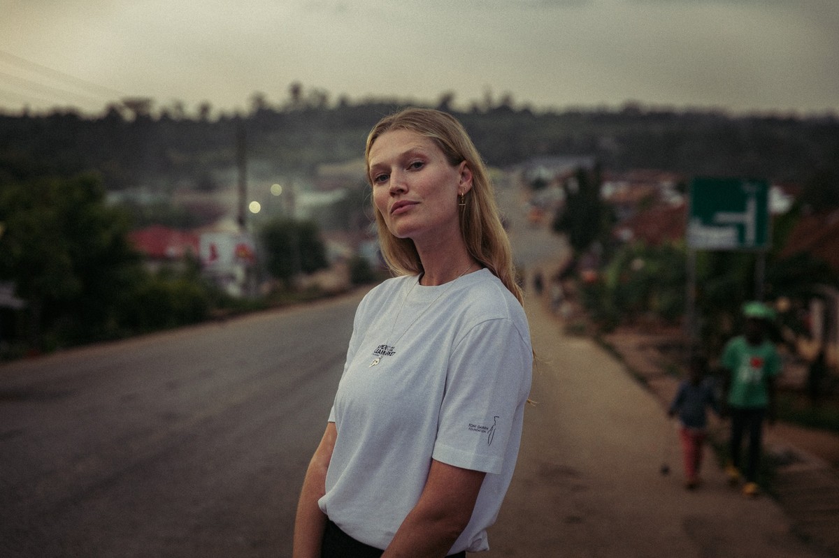 Toni Garrn steht auf einer staubigen Straße in Uganda, im weißen T-Shirt mit dem Aufdruck Super Flea Market, im Hintergrund zwei Kinder, Häuser und Bäume im Abendlicht