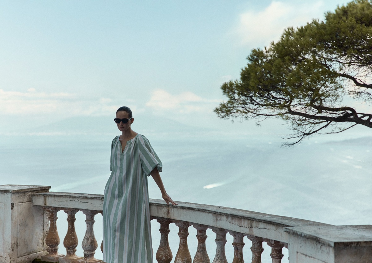 Frau steht auf einer Terrasse mit Blick auf das Meer und trägt ein weit geschnittenes Kleid aus gestreifter Bio-Baumwolle in Grün und Weiß