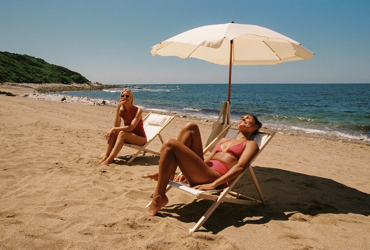 Zwei Frauen entspannen an einem Sandstrand unter einem weißen Sonnenschirm. Im Hintergrund erstrecken sich Meer, Küstenlinie und blauer Himmel.