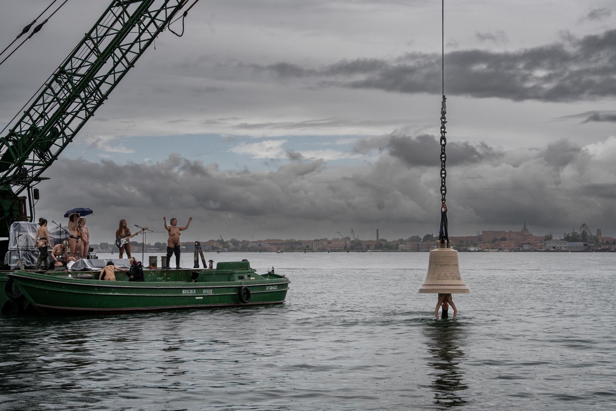 Eine menschengroße Messingglocke wird vor einer schwimmenden Open-Air-Tribüne aus der Lagune von Venedig geborgen. Als menschlicher Klöppel hängt eine nackter Frau aus der Glocke. Auf einem Ponton stehen nackte Musikerinnen mit ihren Instrumenten BU: