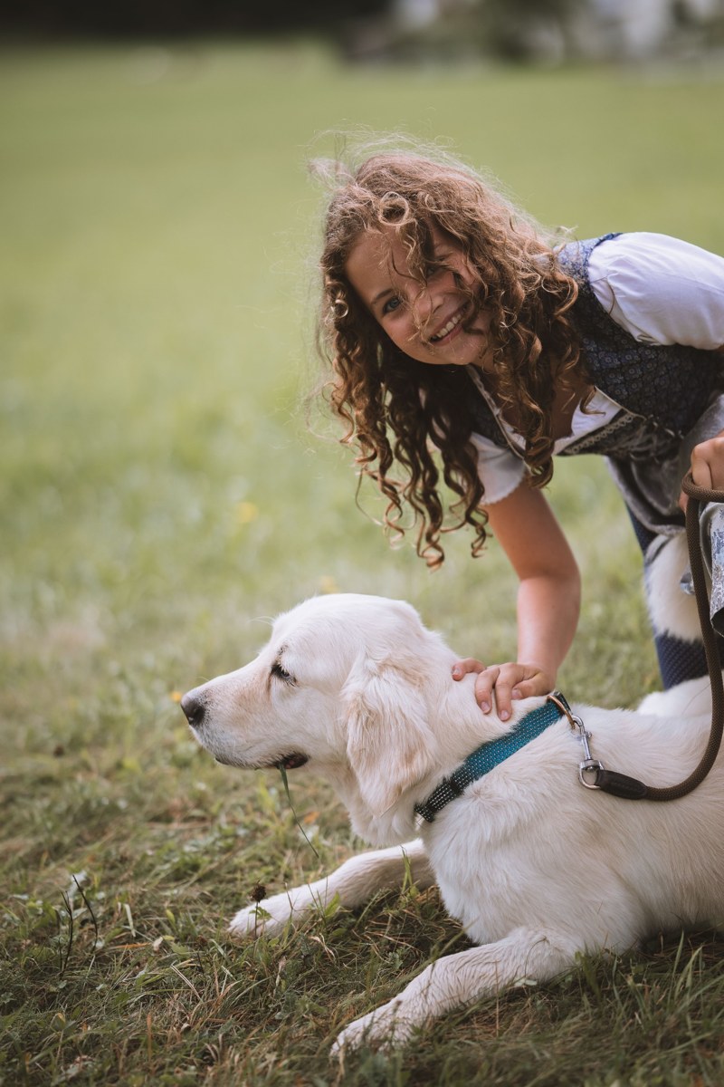 substantial-times-garberhof-03 Mädchen streichelt einen weißen Hund auf einer Bergwiese vor Waldkulisse in Südtirol
