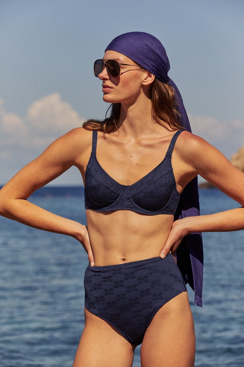 Frau am Meer trägt einen marineblauen Bikini mit hohem Bund und feinem Jacquardmuster. Auf dem Kopf trägt sie ein passendes Tuch und eine Sonnenbrille